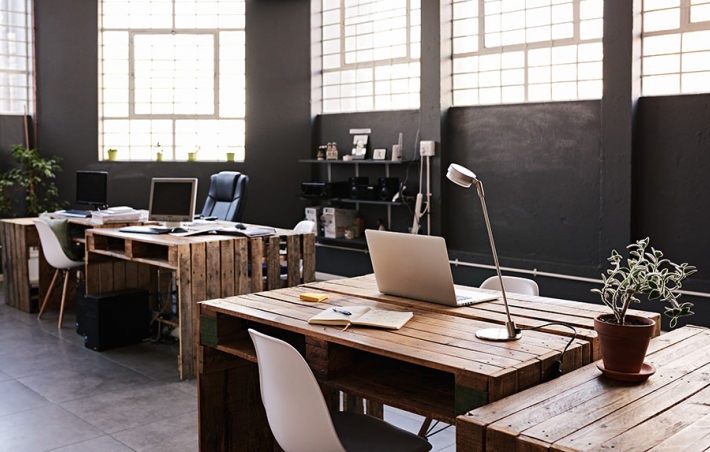 photo of a dark-themed empty office with high ceilings and many high windows. the desks are light wood and littered with laptops, airplants and desk lights