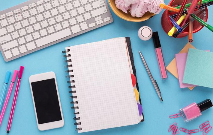 bird's eye view of a blue desk with a notebook, keyboard, phone and some other various desk supplies