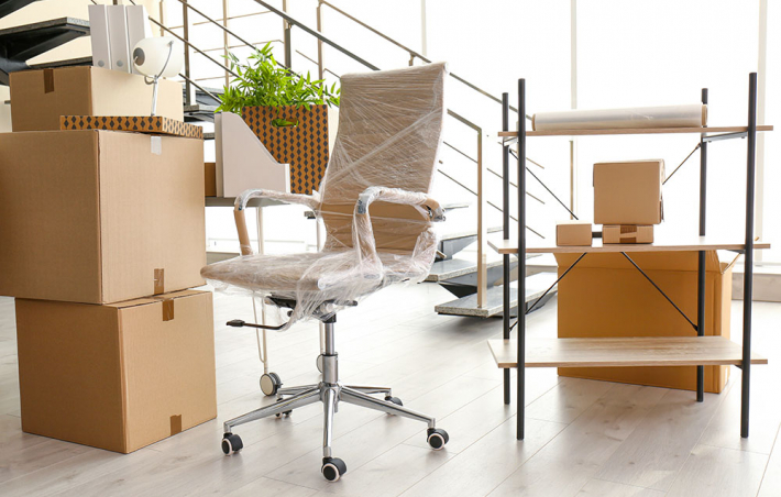 photo of a brightly lit room with unopened boxes, a wrapped office chair, and a small book stand in it. there's a staircase behind the listed items