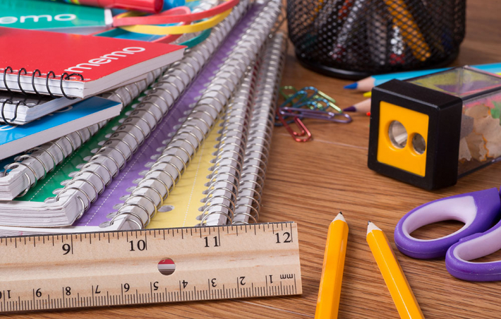 photo of a close-up of a desk littered with a stack of notebooks to the left, a ruler in front of those, and some pencils, a pair of scissors, and a pencil sharpener on the right
